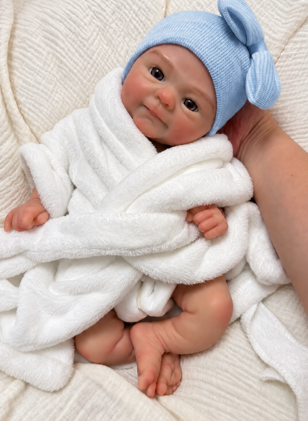 A realistic baby doll or prop baby wrapped in a white plush blanket, wearing a light blue knit hat with a bow, resting on a textured cream blanket with a hand holding it steady.


